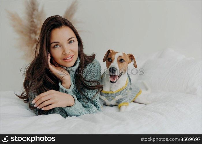 Horizontal shot of pleasant looking tender woman spends free time with favorite pet, looks directly at camera, enjoys domestic atmosphere, cares about dog. People, animals and friendship concept