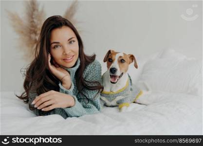 Horizontal shot of pleasant looking tender woman spends free time with favorite pet, looks directly at camera, enjoys domestic atmosphere, cares about dog. People, animals and friendship concept
