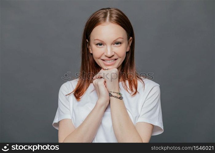 Horizontal shot of pleased beautiful woman wears casual white t shirt, keeps hands under chin, smiles gently, being in good mood, poses against dark grey studio background. Positive emotions