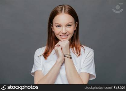 Horizontal shot of pleased beautiful woman wears casual white t shirt, keeps hands under chin, smiles gently, being in good mood, poses against dark grey studio background. Positive emotions