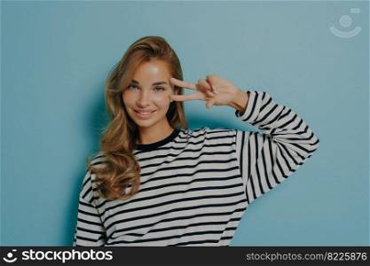 Horizontal shot of pleased lovely young woman with long hair shows peace sign over eye smiles gently feels self confident stands against blue background in casual clothes. Body language concept. Horizontal shot of pleased lovely young woman with long hair shows peace sign over eye