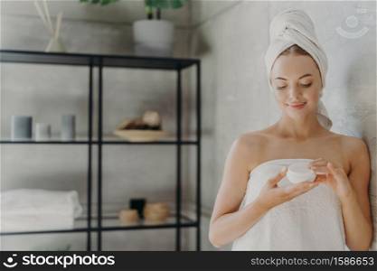 Horizontal shot of pleased woman with healthy skin applies body cream, uses cosmetic product for caring of herself, poses in bathroom after spa bath, undergoes hygiene procedure. Cosmetology concept