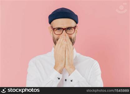 Horizontal shot of positive glad male expresses positive emotions, covers mouth with both hands, giggles silently, wears headgear, white shirt and optical eyewear, isolated over pink background.