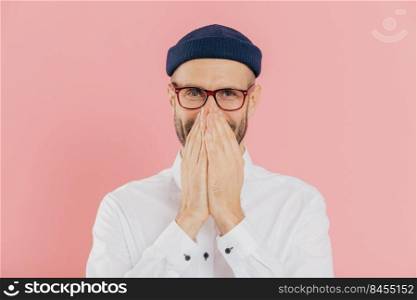 Horizontal shot of positive glad male expresses positive emotions, covers mouth with both hands, giggles silently, wears headgear, white shirt and optical eyewear, isolated over pink background.