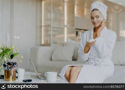 Horizontal shot of pretty Caucasian woman applies facial cream looks thoughtfully aside holds bar of cosmetic product dressed in bathrobe and towel sits on comfortable sofa over home interior