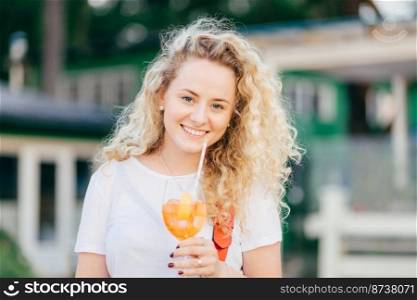 Horizontal shot of pretty curly young female with charming smile, holds glass of fresh orange cocktail, has stroll outdoor, stands against blurred background. People, walk and spare time concept