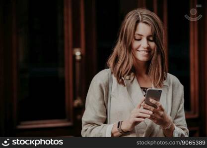 Horizontal shot of pretty cute brunette female model shares multimedia files via cell phone, dressed in elegant clothes, reads positive news in internet website, connected to wireless internet.