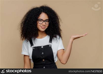 Horizontal shot of pretty dark skinned curly girl raises palm, holds empty space, wears transparent glasses, white t shirt and overalls, isolated over brown background, advertises some object