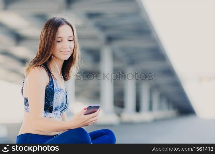 Horizontal shot of pretty lovely woman with pleasant appearance, listens radio, holds modern cell phone, uses earphones and free wifi connection, dressed in sportsclothes. Technology concept