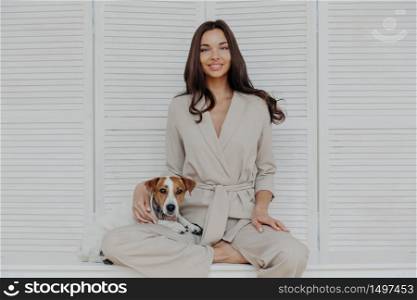 Horizontal shot of pretty relaxed woman dressed in beige costume, plays with favorite dog at home, being in love with domestic pet, expresses affection, being in good mood. Family, lifestyle concept