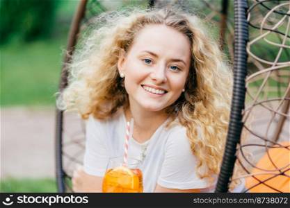 Horizontal shot of pretty smiling European female has wavy bushy light hair, holds fresh cocktail, sits on hanging chair, breathes fresh air. Positive emotions, recreation and people concept