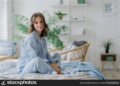 Horizontal shot of pretty woman sits in lotus pose on bed, dressed in casual outfit, listens pleasant melody in earphones, sits against cozy interior. People, domestic atmosphere and relaxation