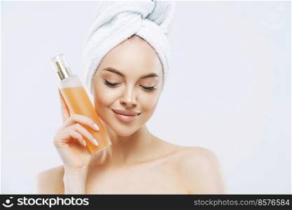 Horizontal shot of pretty woman with healthy skin, natural makeup, uses perfume spray, has pleased expression, natural beauty, looks down, wears bath towel on head, isolated over white background