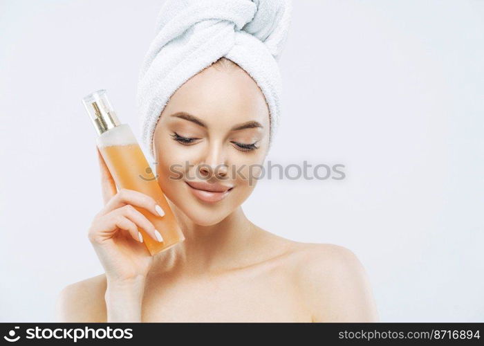 Horizontal shot of pretty woman with healthy skin, natural makeup, uses perfume spray, has pleased expression, natural beauty, looks down, wears bath towel on head, isolated over white background