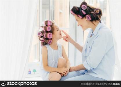Horizontal shot of pretty young mother and daughter with curlers on hair, being in good mood, enjoy time at home, prepare for holiday. Happy family concept. Beauty, children and fashion concept