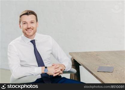 Horizontal shot of prosperous male entrepreneur in formal clothes, holds hands together, has positive expression, sits at wooden table with touch pad, waits for business partners to have meeting