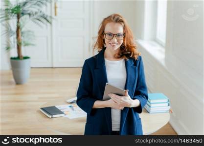 Horizontal shot of proud CEO checks email on digital touchpad, looks at camera with satisfied expression, wears spectacles, stands in modern office, enjoys online communication on modern device