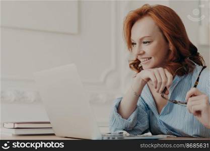 Horizontal shot of redhead European woman poses at home office, focused at laptop screen, holds spectacles, watches video on computer, enjoys good news, wears shirt, enjoys distance learning
