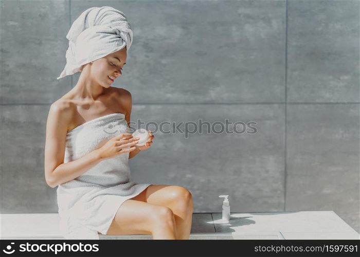 Horizontal shot of relaxed beautiful womam applies body lotion and nourishing cream after shower, poses in bathroom against grey wall, sits wrapped in towel, undergoes beauty treatments. Spa concept