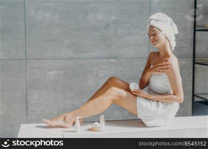 Horizontal shot of relaxed beautiful young woman spreads cream on shoulder, uses cosmetic and takes care of her skin has slender legs wrapped in bath towel enjoys spa procedures poses with closed eyes