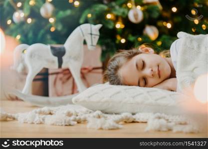 Horizontal shot of relaxed girl covered with white sweater, has pleasant dreams, lies under decorated Christmas tree, toy rocker and gift boxes around. Kid at home. Cozy holiday. Domestic atmosphere