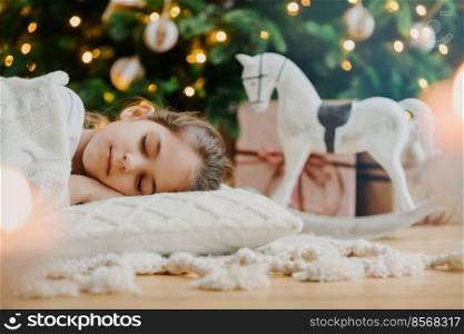 Horizontal shot of relaxed girl covered with white sweater, has pleasant dreams, lies under decorated Christmas tree, toy rocker and gift boxes around. Kid at home. Cozy holiday. Domestic atmosphere