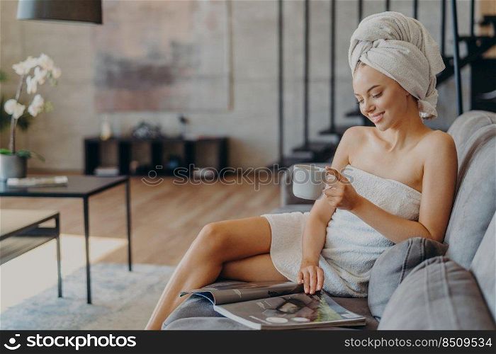 Horizontal shot of relaxed satisfied woman has healthy skin reads magazine poses with cup of coffee or tea at couch wrapped in soft towels enjoys spending time at home. Refreshment leisure spa