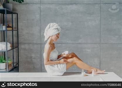 Horizontal shot of relaxed young European woman applies moisturizing body cream on legs, wrapped in bath towel, has tender smile, healthy refreshed skin after taking bath, poses in cozy bathroom.