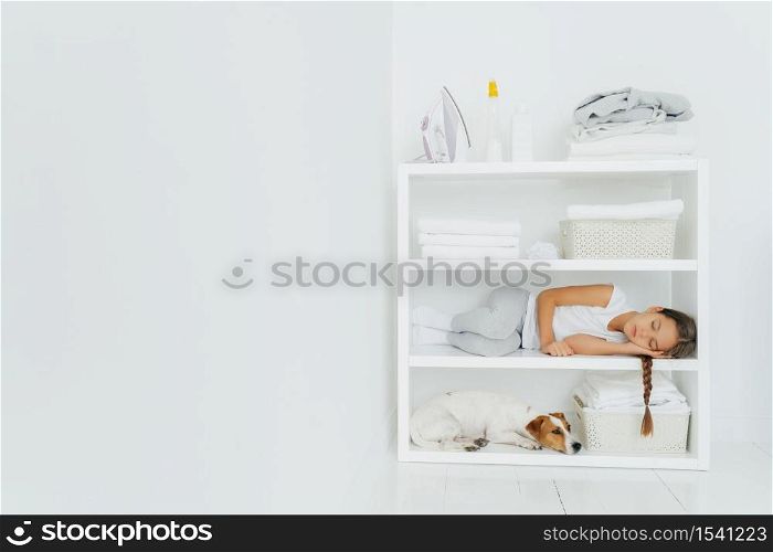 Horizontal shot of restful girl lies on console with dog, rests after folding towels in washing room, fall asleep after domestic work, white wall, copy space on left side for your promotional content