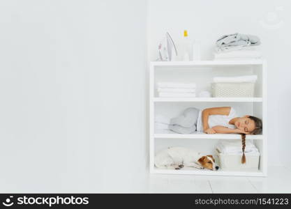 Horizontal shot of restful girl lies on console with dog, rests after folding towels in washing room, fall asleep after domestic work, white wall, copy space on left side for your promotional content