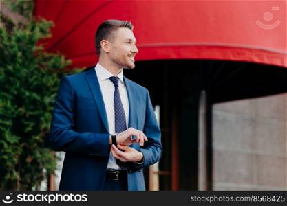 Horizontal shot of satisfied businessman in formal clothing, looks at watch, waits for partner near cafe, going to discuss details of collaboration, has glad expression, poses outside. People and job