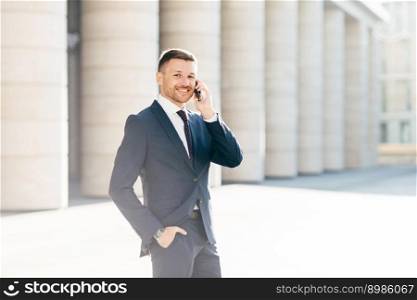 Horizontal shot of satisfied male with positive look solves banking problems while calls to operator via smart phone, wears elegant black suit, poses putdoor in urban setting. Communication concept