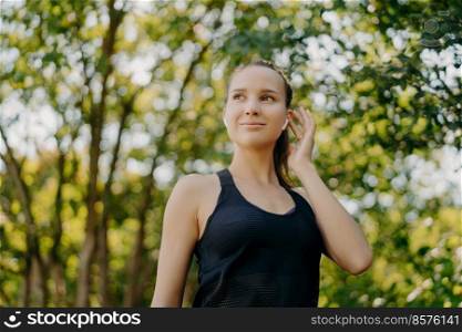 Horizontal shot of satisfied young woman keeps hand on earbud listens favorite music looks away has satisfied face expression dressed in active wear enjoys summer morning outdoors has cardio workout