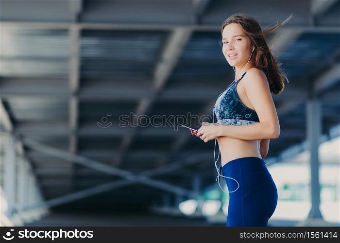 Horizontal shot of slim athletic female with perfect figure, dressed in casual top and leggings, holds modern cell phone, listens popular songs in earphones, poses outdoor. People and rest concept