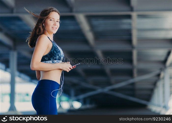 Horizontal shot of slim athletic female with perfect figure, dressed in casual top and leggings, holds modern cell phone, listens popular songs in earphones, poses outdoor. People and rest concept