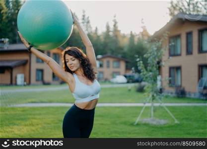 Horizontal shot of slim brunette Caucasian woman practices yoga with fitness ball, exercises outdoor, has gymnastic training, dressed in active wear, poses on green lawn with houses in background