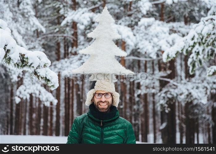 Horizontal shot of smiling male model holds fir tree on head, stands outdor in winter forest, enjoys calmness and tranquil atmosphere. Caucasian unshaven man with cheerful expression