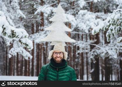 Horizontal shot of smiling male model holds fir tree on head, stands outdor in winter forest, enjoys calmness and tranquil atmosphere. Caucasian unshaven man with cheerful expression