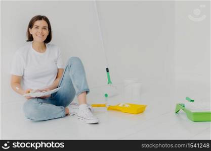 Horizontal shot of smiling woman designer chooses best color for apartment, holds palette, dressed in casual clothes, paints walls, renovates her home, thinks about creative ideas to decorate room
