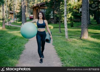 Horizontal shot of sportswoman wears cropped top and leggings, carries fitness ball and rolled up karemat, walk on road around trees and green grass, leads healthy lifestyle. Sport equipment