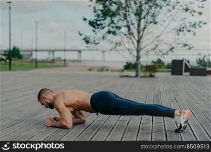 Horizontal shot of sporty bearded man stands in plank pose, practices yoga outdoor and dressed in active wear, breathes fresh air. Sport, fitness and healthy lifestyle concept. Training workout