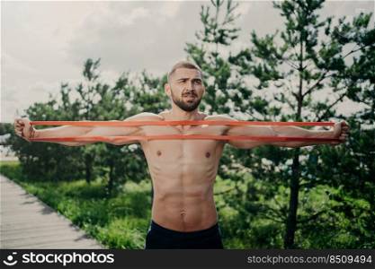 Horizontal shot of sporty man with bare chest exercises with elastic expander, gains for strong muscular body, has thick bristle, poses outside. Bodybuilder stretches band stands near green trees