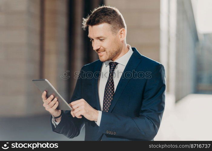 Horizontal shot of successful male office worker dressed in elegant suit, holds modern digital tablet, updates website with financial or business news. Happy trader with modern electronic device
