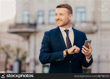 Horizontal shot of successful unshaven male CEO looks for interesting multimedia files on smart phone, downloads or updates new programs, stands against blurred city background, looks happily aside