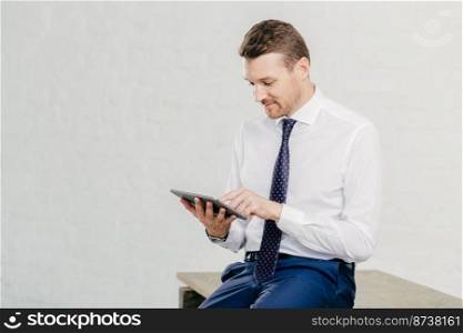 Horizontal shot of successful young businessman updates profile on business webblog uses application on tablet computer, wears elegant clothes, isolated over white background in office building
