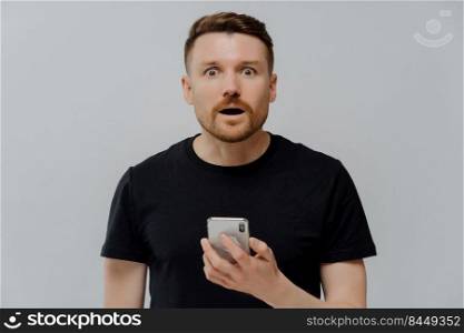 Horizontal shot of surprised startled bearded adult man holds mobile phone reacts on shocking news dressed in casual black t shirt uses modern cellular for online communication poses indoor.