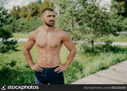 Horizontal shot of thoughtful shirtless sportsman keeps hands on waist, looks away demonstrates his muscular body, poses over nature background. Active bodybuilder goes in for sport, has daily workout