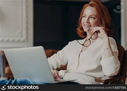 Horizontal shot of thoughtful young redhead woman concentrated somewhere, works on freelance project, holds spectacles, keeps laptop computer on knees, enjoys online job, creats new publication