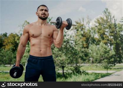 Horizontal shot of topless fitness unshaven European man has muscular body, raises barbells, wears shorts, demonstrates strong arms, has workout outdoor in park. Sportsman makes weightlifting