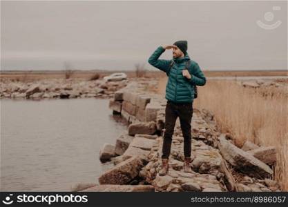 Horizontal shot of unshaven male traveler dressed in warm jacket, hat and shoes, keeps hand near forehead, looks attentively into distance, admires landscapes, stands near lake, carries rucksack